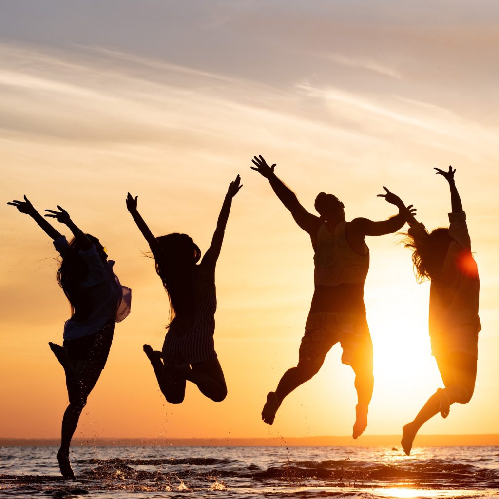 four teens are silouetted by the setting sun as they jump above the waves at the beach with their hands in the air